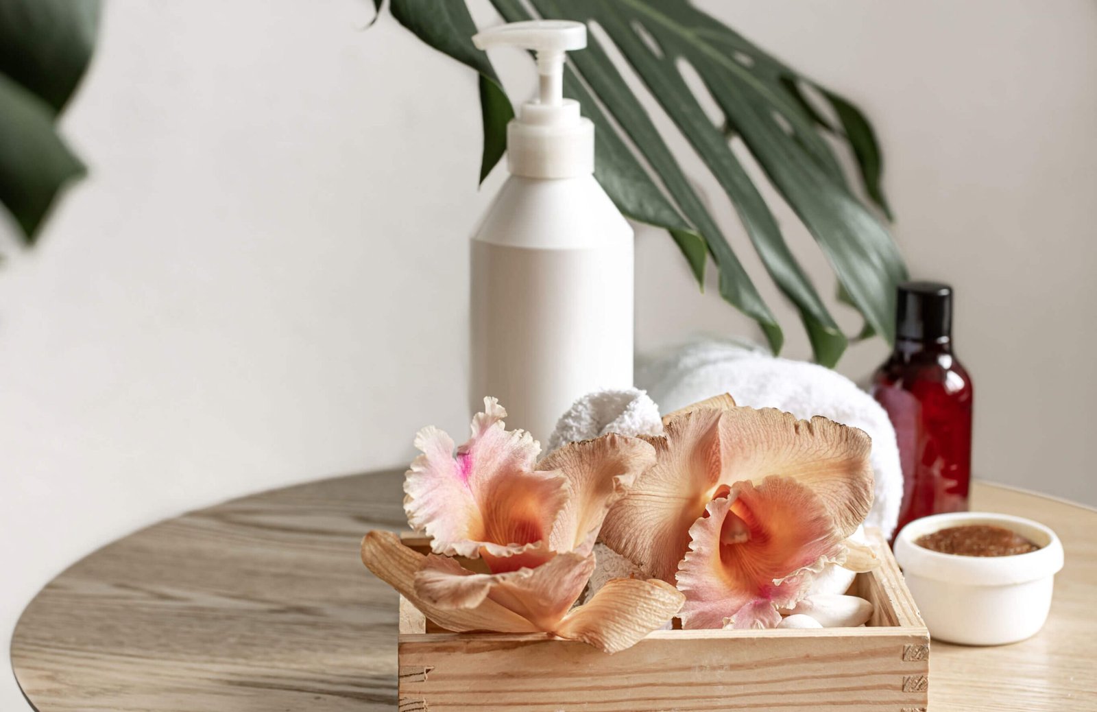 A wooden tray with peach-colored artificial orchids,Natural beauty products, towels, a white lotion bottle, a small red bottle, and a bowl of scrub on a light wooden table with a green leaf in the background.
