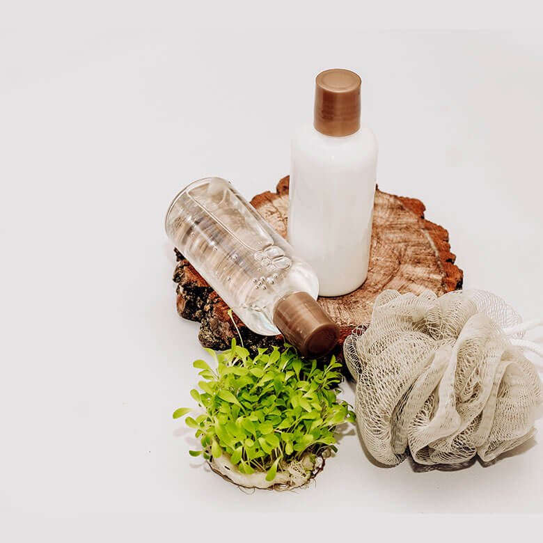 Two bottles of skincare products, one clear and one white, are placed on a wooden slice, next to a green plant and a beige bath pouf, against a white background.