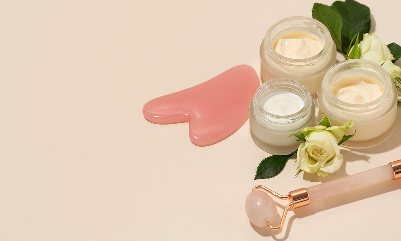 Three open jars of face cream, a pink gua sha tool, a jade roller, and fresh white roses on a light beige background.