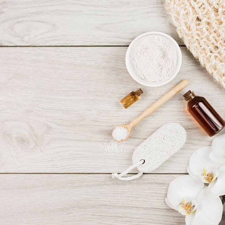 Flat lay of spa items on a light wooden surface, including a bowl of powder, small bottles of oil, a wooden spoon with salt, a pumice stone, a knit cloth, and white orchid flowers in the corner.