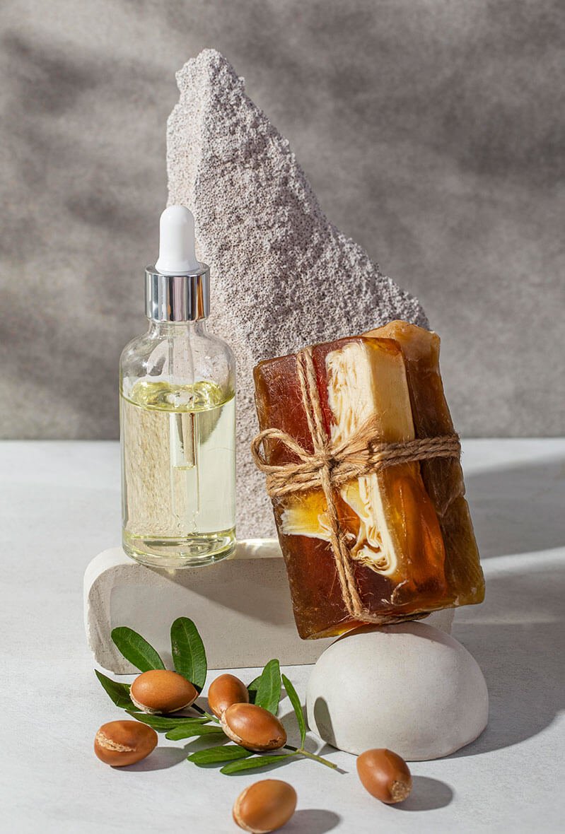 A glass dropper bottle of oil, a tied bar of handmade soap, argan nuts, and green leaves are arranged on white stones with a textured stone backdrop. The scene is bright and minimalist.